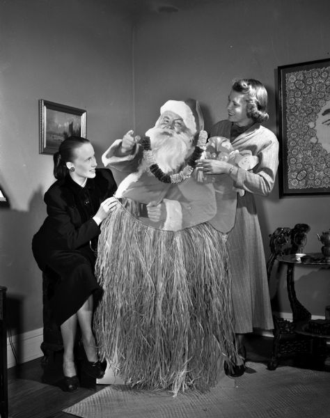 Naval Officers and Wives Club annual Christmas dinner dance featuring a Hawaiian Christmas theme, in the Crystal Ballroom of the Hotel Loraine. Mrs. Robert (Janet) Huegel, 1010 Rutledge Street, and Mrs. Charles (Marcella) Route 3, decorate a cardboard Santa Claus with a grass skirt.