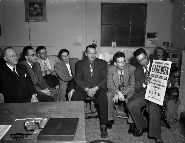 Seven members of the Madison CARE committee looking at a poster  about CARE Week. Left to right are Rev. W.B. Waltmire, Madison Council of Churches; Rabbi Max Ticktin of the Jewish clergy; Fred Brussow, CIO council representative; Mrs. L.E. (Edith) Isaksen, Madison PTA council; Harold Haak, Madison Federation of Labor; Al Reindle, Milwaukee, Wisconsin CARE committee; and Peter J. Slifen, general chairman of the Madison committee. In the rear at right is Janice Walton, Milwaukee CARE committee member.