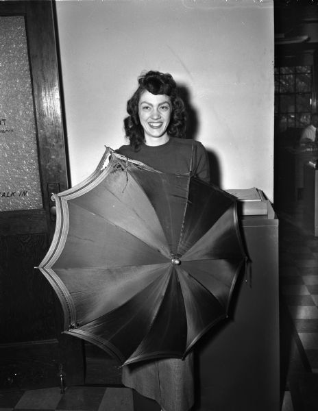 Mrs. Marian M. (Mary) Hammang, secretary in the office of the University of Wisconsin buildings and grounds, posing with an umbrella that was blown by high winds into a high voltage line, short-circuiting the power and causing a dim-out over Madison's west and south sides. The umbrella belonged to Betty Solum of Baraboo, a freshman in home economics at the university.