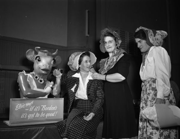 Members of the Madison Who's New Club at a barn dance at the YWCA gymnasium. Three women wearing bonnets pose next to a statue of Borden's "Elsie the Cow." They are, left to right: Mrs. Arlo Miller, 1820 Rutledge Street; Mrs. John I. Cole, 3227 Shore Acres Road and Mrs. (Grace) Paul Fakler, 1347 Jenifer Street.