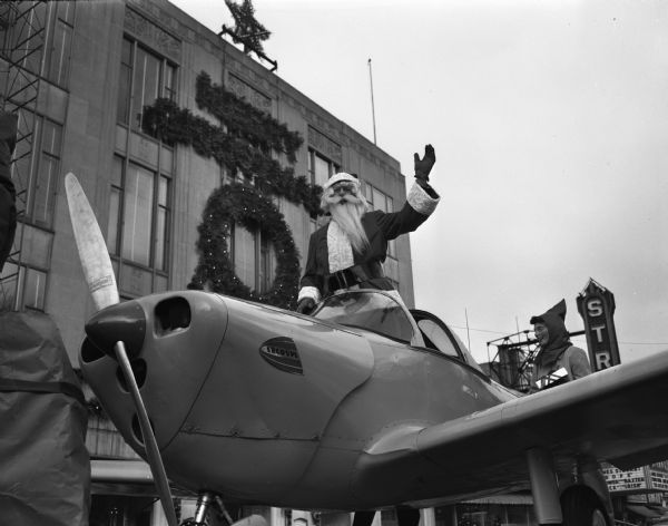 In front of Manchester's Department Store, Santa Claus waves to the children from his yellow Ercoupe airplane as he rides in a parade sponsored by the Madison Business Association and the Junior Chamber of Commerce.