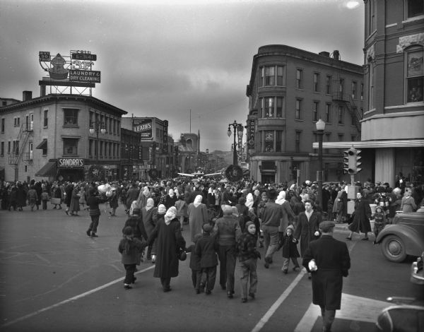 Crowd at the corner of State, West Mifflin and North Carroll Streets watching the Santa Parade, sponsored by the Madison Business Association and the Junior Chamber of Commerce, come up State Street toward the Square. In the middle background Santa Claus can be seen waving to the crowd from his yellow Ercoupe airplane.