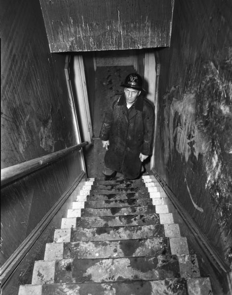 Fireman Orrin Zebarth is shown at the foot of the narrow, steep front stairs which occupants were forced to use when flames cut off escape routes at the rear of the tenement-type house, which is located behind the  city garage. The fire at 15 South Bedford Street killed Florence Dinger and three of her children.