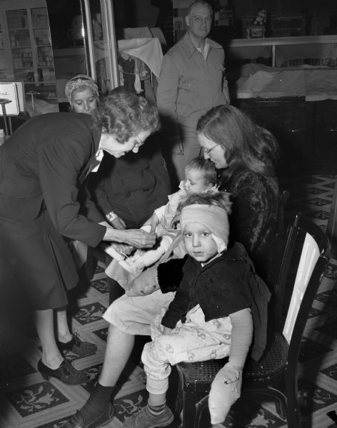 Bertha Atkinson, Baron Brothers Department Store buyer, fits a bootie on Norma Widener, 11-months old, being held by her mother Valera Widener. In the foreground is Lyall Widener, 3, who like his mother was severely burned. Behind Mrs. Atkinson is Josephine Coleman, whose son, John Collins Coleman, 11, was among the children helped by the Empty Stocking Club following a fire in the tenement-type house at 15 South Bedford Street, which killed Florence Dinger and three of her children.