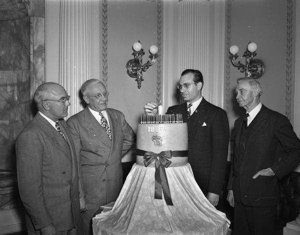 Four men standing around a large block of cheese decorated with candles and a ribbon. Governor Oscar Rennebohm second from left. Engraved on the cheese are the words "100th" and within an outline of the state, "State Department of Agriculture Wisconsin State Brand 163-B."