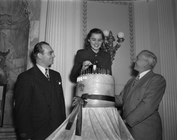 Governor Rennebohm, right, with unidentified man and woman standing around a large block of cheese decorated with candles and a ribbon.  Engraved on the cheese are the words "100th" and within an outline of the state, "State Department of Agriculture Wisconsin State Brand Cheese 163-B." The woman lighting the candles may be Marjean Czerwinski, the 1951 Alice in Dairyland.