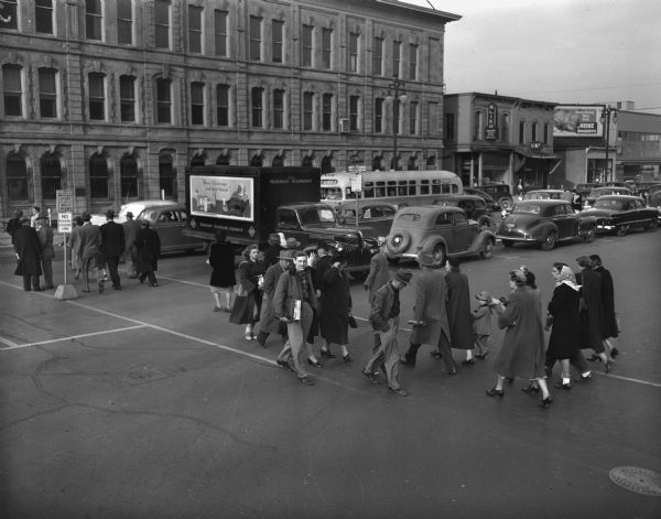 Elevated view of street scene at East Washington Avenue and Pinckney streets with pedestrians crossing the street. Advertisement on the side of a parked Railroad Express Agency Truck has Santa Claus saying "Merry Christmas and Good Health."