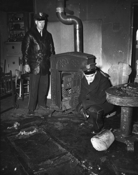 Police officers Frank Eisele and Robert O'Neil examining the explosion-wrecked stove and the kerosene can from which resident Walter Alburn poured kerosene on the embers in the wood-burning stove, resulting in the explosion and fire in the apartment of the Truax barracks.