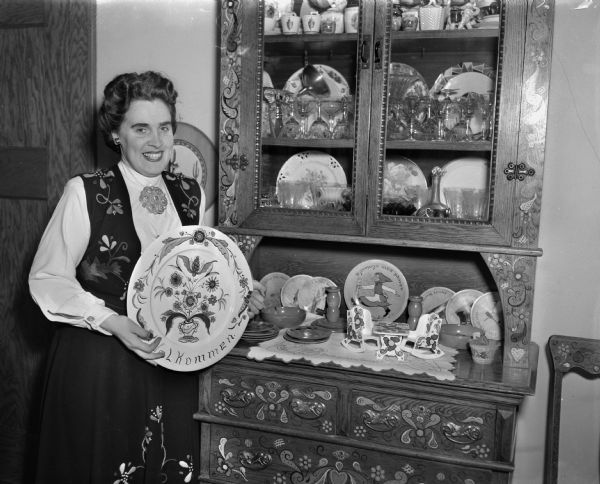 Mrs. Celin Furu is shown in Norwegian costume displaying her rosemaled painted platters and sideboard cabinet. She was a wartime resident of Bergen, Norway when it was bombed 57 times in the month of June, 1940.