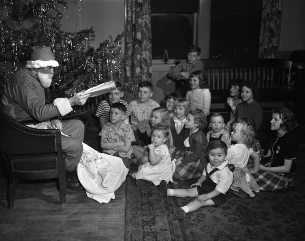 Children of Milwaukee Railroad employees sitting near a Christmas tree where a man sitting in a chair is dressed as Santa Claus. They are at the Milwaukee Railroad Women's Club, 640 West Washington Avenue.
