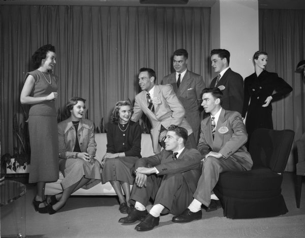 Three female models that took part in a fashion show for the Wisconsin All-State Football Squad during a dinner at Manchester's Madison Room, shown with five of the all-staters. Standing is Marjorie Groat of St. Paul, Minnesota who was featured in national publication ads. Seated are Barbara Mathys (left), a Madison East High School graduate now attending school in the east; and Phillis Berg, a Wisconsin High School senior. The football players are Ronald "Red"  Walker, Durand (seated on floor); Rollie Bestor, Green Bay West (on back of the divan); Joe Pelikan, Edgerton (seated on chair); and Bob Kennedy, Rhinelander, and Tom O'Brien, Wauwatosa (standing).