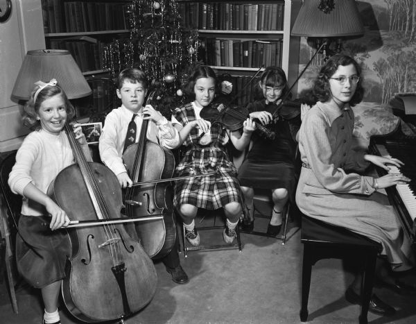 Shorewood Hills music students posed with their string instruments at a Christmas caroling party in the home of Mrs. Keys (Freda) Winterble, 901 University Bay Drive, Shorewood Hills. The students are, left to right: Harriet Irwin, David Neal, Barbara Bullington, and Priscilla Butts. A woman sits at a piano on the right.