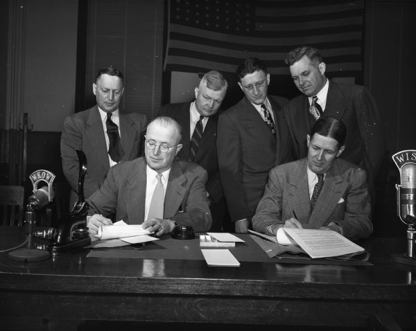 Dane County Board Chairman Paul A. Robinson, left, and Madison City Manager Leonard G. Howell, right, signing contracts which officially join Madison and Dane County in the construction of a new dual government building. Standing behind are, left to right: County Clerk Austin N. Johnson; Darwin Bruns, Chairman of the county board city county building committee; City Clerk A.W. Baresis; and City Council President Henry Reynolds. On the table are two radio broadcasting microphones.