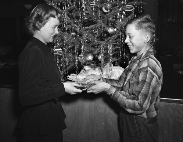 Dorothy Blank receiving donated money for the Empty Stocking Club from newspaper carrier boy Gordon Schaaf in front of a Christmas tree.