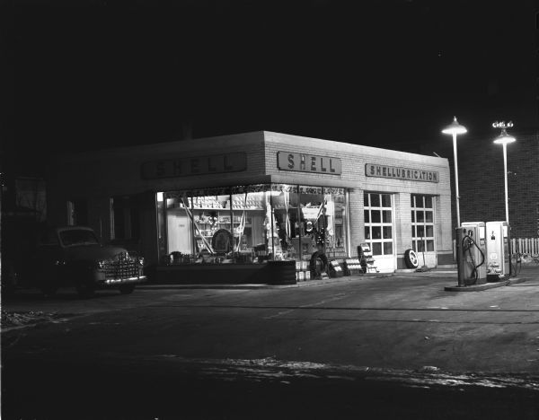 Night view of Shell Gas Station at 1200 East Washington Avenue, operated by Harold W. Risberg. There is a holiday display inside the station behind plate glass windows, including a cardboard easel-back display of Santa.