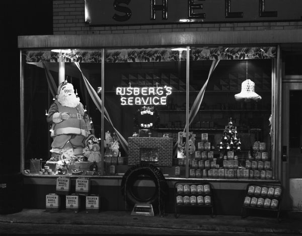 Night view of display window of gas station featuring a cardboard easel-back display of Santa Claus, and an artificial fireplace among other decorations, at the Harold W. Risberg Shell gas station at 1200 East Washington Avenue.