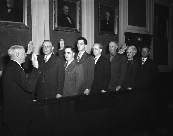 Group portrait of seven Dane county officials--five new and two reelected--shown taking the oath of office before Circuit Judge Herman W. Satchjen at far left. Pictured left to right: Marvin Smithback, county treasurer; Miles Riley, Jr., register of deeds; Keith A. Schwartz, county clerk; Robert Arthur, district attorney; Andrew Dahlen, surveyor; Myrtle Hansen, clerk of county; and Herman Kerl, sheriff.