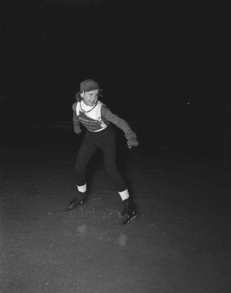 Portrait of Patsy Gibson, wearing a Wisconsin Felton Sporting Goods shirt, poised to begin an ice speed skating race. Patsy, age 14, daughter of Mr. & Mrs. Ed Gibson, 609 South Shore Drive, placed second in the Junior Girls class in both the 220 and 440 yard dashes of the Silver Skates meet of the Wisconsin Skating Association in Milwaukee.  She is the Madison juvenile champion.