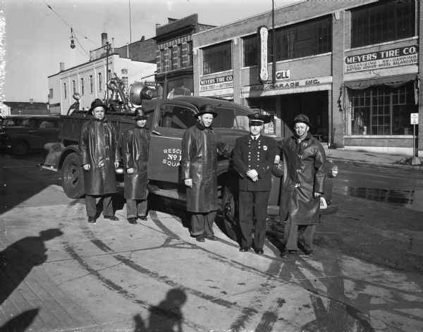 Five fire fighters, Kendall Niebuhr, William J. Sullivan, John G. Randall, Chief Edward J. Page, and Gill S. Stone, pose with newly customized rescue truck in front of Central Fire Station #1, 18-20 South Webster Street. Businesses across the street are: Gill Garage, 13-15 South Webster Street, and Moose Club, 9 South Webster Street.  The truck was purchased from Truax Field for $200.