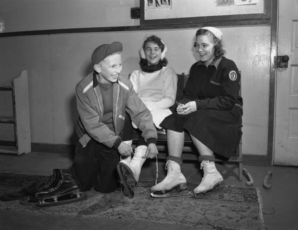 Arthur Stehr, son of Dr. and Mrs. A.C. Stehr, 3502 Blackhawk Drive, puts on the skates of Nancy Blume, daughter of Mr. and Mrs. Norman H. Blume, Shorewood Hills, while Francesca Axley, daughter of Mr. and Mrs. Ralph E. Axley, 3515 Sunset Dive, looks on at the Madison Figure Skating Club's new indoor skating arena at Truax Field.