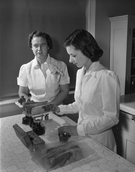 Dorothy Hussemann, associate professor of home economics at the University of Wisconsin, (left) addressing her class on the technique of weighing meat. At right, student Louise Borden, 1922 Adams Street, demonstrates the process. Professor Hussemann was recently chosen to judge the <i>Wisconsin State Journal's</i> first recipe contest.