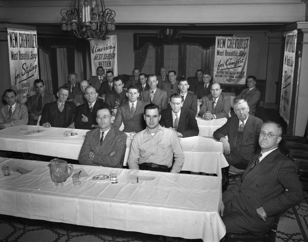 Group of salesmen for the Chevrolet Company at a sales meeting. Advertising banners are visible on the walls.