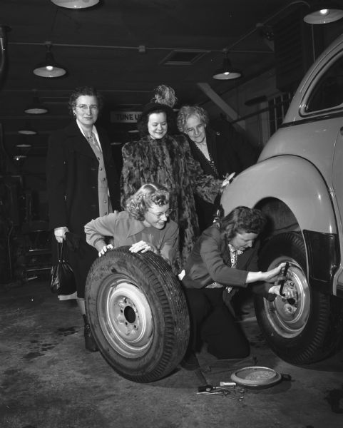 Charlotte Holmes (with tire) and Mrs. Lee (Marjorie) Edwards (with wrench) change a tire during a class session at Madison Vocational School's first post-war driving course. Looking on, left to right, are: Mrs. J.H. (Isophene) Duff, Mrs. George C. (Janet) Hank, and Mrs. Frieda Taplick.