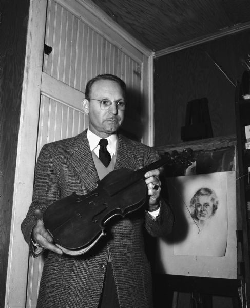 Paul C. Bennett, musical instrument repairman, holding a violin made in 1721 which belongs to Mrs. D.P. Wheeler and may be a Stradivarius. Behind him on an easel is displayed a drawing of a woman.