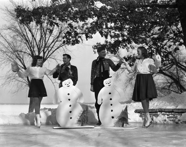 Four University of Wisconsin students wearing figure skating costumes are shown posing in front of Lake Mendota where they performed a show on Memorial Union Terrace. Left to right, are: Anita Schmidt, holding a muff, Milwaukee; Michael Bartelme, Superior; Richard Symmes, Pasadena, California; Anne Blakeley, also holding a muff, Chester, Pennsylvania.