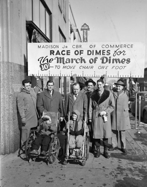 Two children in wheelchairs, Jean Bokina, left, daughter of Mr. and Mrs. Edward Bokina and right, Steven Butts, son of Mr. and Mrs. F. Freeman Butts, with members of the Junior Chamber of Commerce, left to right: Dr. Fritz Krohn, Arlie Mucks Jr., Henry Gempler, Charles Swanson, Lee Baron and Bob Smith. This photograph was taken just prior to a race around the Capitol Square. The "race of dimes" is part of  March of Dimes fundraising.