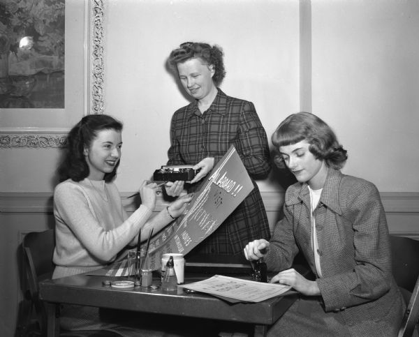 Three women preparing for a YWCA benefit party to send representatives to an YWCA conference. Delores Nelson, left, is painting a poster.  Violet Fisher, center, chairman of the committee, is holding paint jars, and Phyllis Nickerson, right, is numbering admission tickets.