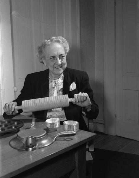 Miss Gladys S. Stillman, judge of the "Wisconsin State Journal's" dessert recipe contest, exhibiting utensils used in baking nationality dishes. Also in the picture are a lefse (rolling pin), a Scandinavian krumkske iron, a munk pan for making Danish aebeleskiver, and small tins for making sand bakkels which are a rich Scandinavian cookie. She is a nutrition specialist in the University of Wisconsin home economics extension service.