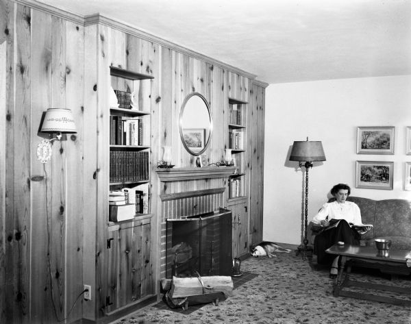 The knotty pine living room in the home of the Edward B.A. Sokoloski family at 874 Woodrow Street. Mrs. Lucille Sokoloski is reading a book, and the family dog, Peppy, is taking a nap in the corner.