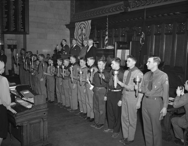 Scouts representing each of the 17 Boy Scout councils of Wisconsin line up with torches inside the Wisconsin State Capitol to open their national campaign to "strengthen the arm of liberty". In the background, left to right, are Harlin Nicholls, Four Lakes council president; Jack Raymond, council commissioner; Gov. Rennebohm; and Chief Justice Rosenberry.
