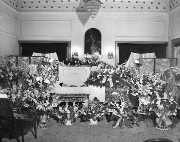 Funeral service of Mrs. Gerald (Cora) Fries, wife of Blooming Grove Constable "Jerry" Fries, showing the open casket in the Gunderson funeral home, 936 Winnebago Street, surrounded by many floral arrangements.