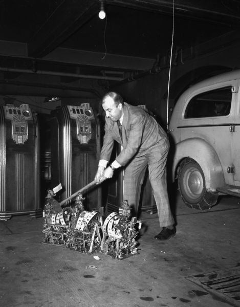 Madison Police Inspector Philip H. Oakey is shown demolishing three "one-armed bandits." They had been stored in the vault of police headquarters since they were confiscated during a gambling raid at the Madison Novelty Company, formerly operated at 606 University Avenue.