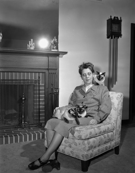 Portrait of Mrs. Westye F. (Mary) Bakke seated before the fireplace in the living room of the Bakke house at 4818 Odana Road (now 4817 Sherwood Road) with her three favorite Siamese cats.