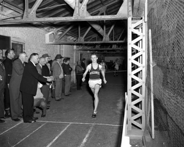 Bob Mansfield, University of Wisconsin track athlete, crosses the finish line as he breaks the annex record for the indoor quarter mile at the University of Wisconsin Athletic Annex. A group of men cheers him on from the sidelines.