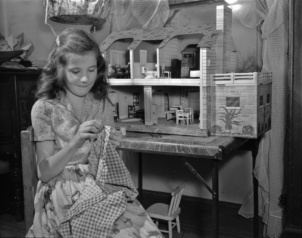 Mary Henshue, age 10, has just become eligible for 4-H. She is shown sewing doll clothes. She is a member of the Silver Badgers 4-H Club. A dollhouse is on the table behind her.