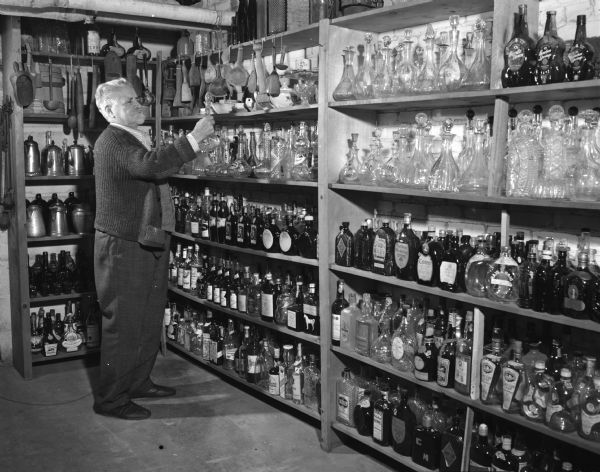 A man is standing next to shelves housing his bottle collection.