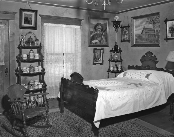 House interior with a Victorian bedroom decor. The room contains a rocking chair, a sleigh bed, and shelves holding decorative steins and other figurines. Framed paintings and a photograph are hanging on the walls.