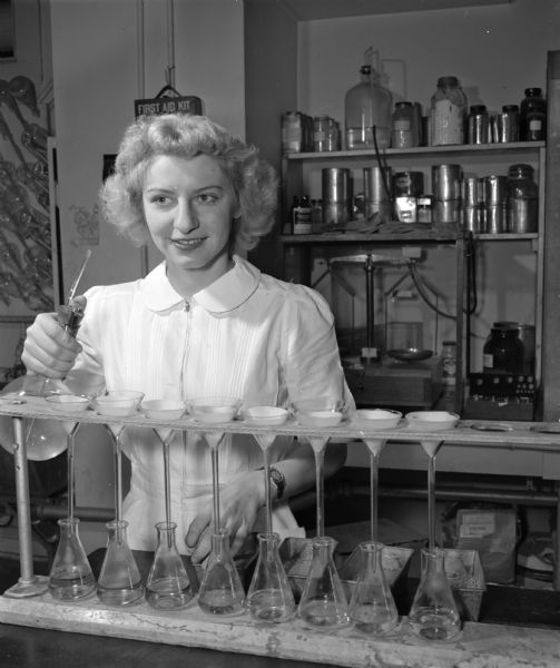 A woman stands near a row of beakers in a chemistry laboratory.  Various jars and other equipment are visible on a shelf behind her.