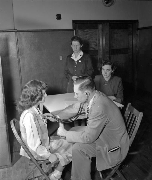 A male doctor examines a young female patient while two women look on.