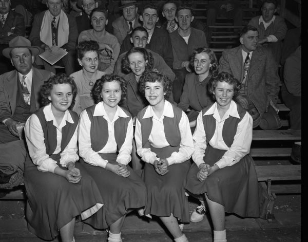 Fans and cheerleaders from Kimberly at the state high school basketball tournament. Cheerleaders in front, left to right, are Laverne Kamps, Joanne Lamers, Bette Vander Velden, and Birdie Kneepkens. Behind the cheerleaders are, left to right, Mrs. Arnold Leaman, wife of the Kimberly coach; Mrs. Robert DuCharme, wife of the assistant coach; and Jean Behling, a member of the State Journal editorial staff and former Kimberly resident. Other fans are sitting in the bleachers behind them.