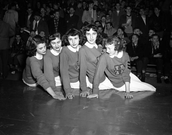 Hurley cheerleaders posing on the floor at the state high School basketball tournament with spectators sitting behind them. Left to right are: Loretta Cortichiate, Gloria Gunnunglo, Jean Ann O'Birto, Rose Marie Pasqualucci, and Joanne Kopack.