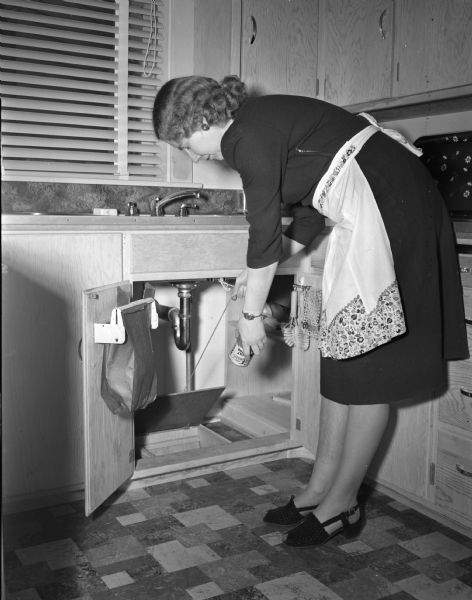 Mrs. Edith Trowbridge, 4202 Mandan Cresent, pulls a ring on a string which opens a trap door to a metal-lined slide to a basement trash box under her sink in her kitchen. L.G. Trowbridge constructed the trash disposal device.
