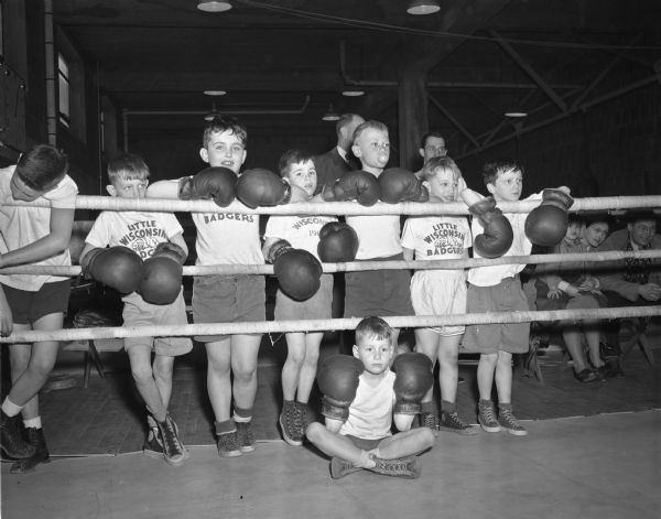 Eight boys watching a boxing match from outside the ring during the Little Badger boxing classes at the University of Wisconsin-Madison Field House. Sitting is Bobby Johnston. Standing, left to right, are an unidentified boy hanging his head, John Halverson, Tim Jones, Johnny Walsh, Jr., Bruce Hasler, Larry Lulling, and Mike Jordan. Coach Johnny Walsh is in the background.