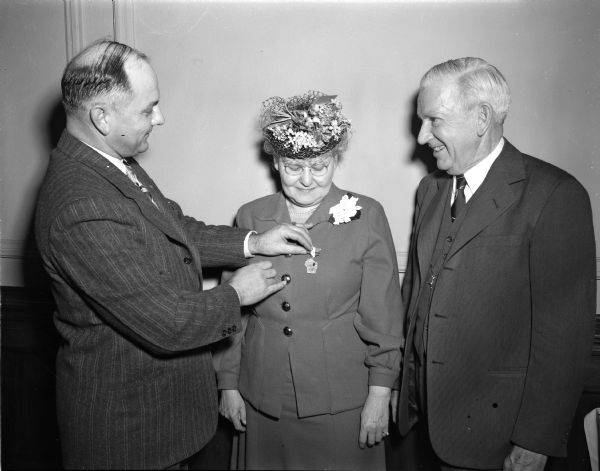 Two men present a medal to an elderly woman wearing a floral hat and corsage on her lapel.