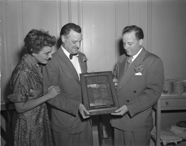 Mautz Paint and Varnish Company salesman, Alfie Ladd (right) presents a bronze plaque inscribed with signatures of the firm's sales force to company president Bernhard Mautz at a 50th birthday party for him at the Madison Club. Mautz's wife Jane is standing on the left.