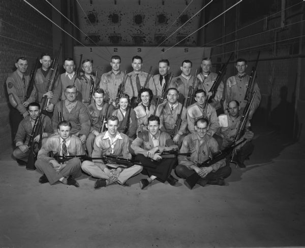 Twenty-two men and women pose with their rifles in a indoor rifle shooting range.